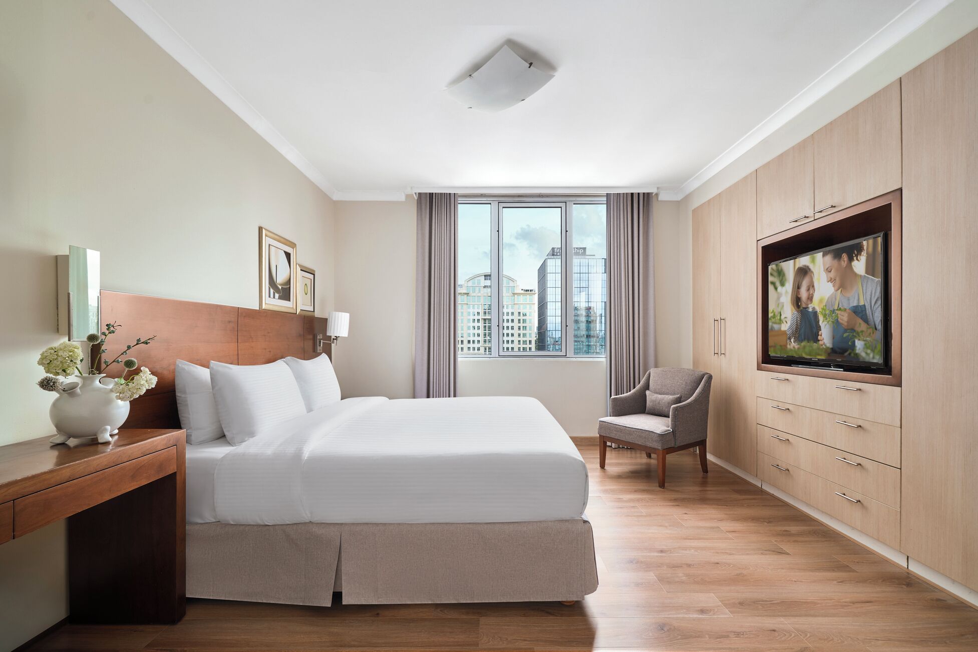A hotel room with a bed, a TV, a chair, a desk, and a window with a city view.
