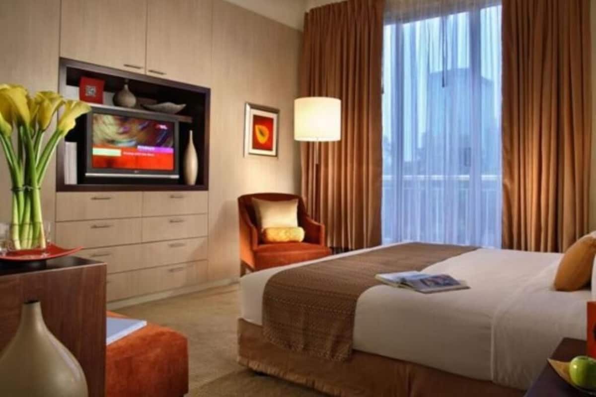 A hotel room with a bed, a TV, a chair, a lamp, and a window with curtains.