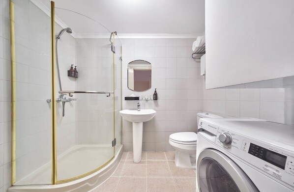 Un baño moderno con ducha envuelta en vidrio, lavamanos pedestal, inodoro y lavadora de carga frontal.