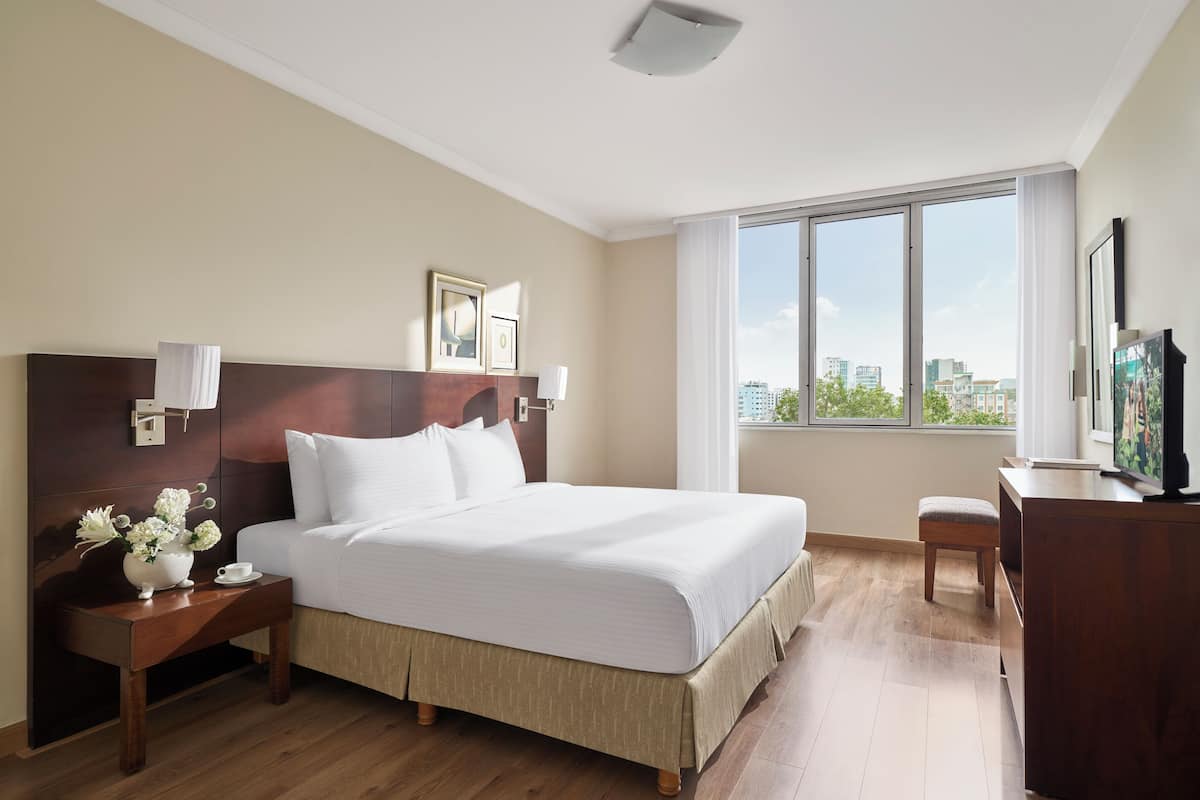 A hotel room with a large bed, a nightstand with a vase of flowers, a desk with a television, and a window with a city view.