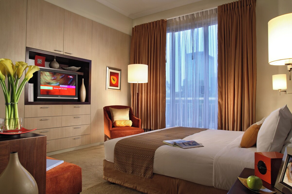 A hotel room with a bed, a TV, a chair, a desk with a lamp, and a window with curtains.