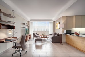 Moderni hotellihuone, jossa on sohva, nojatuoli, työpöytä ja suuret ikkunat, joista avautuu näkymä kaupunkiin.