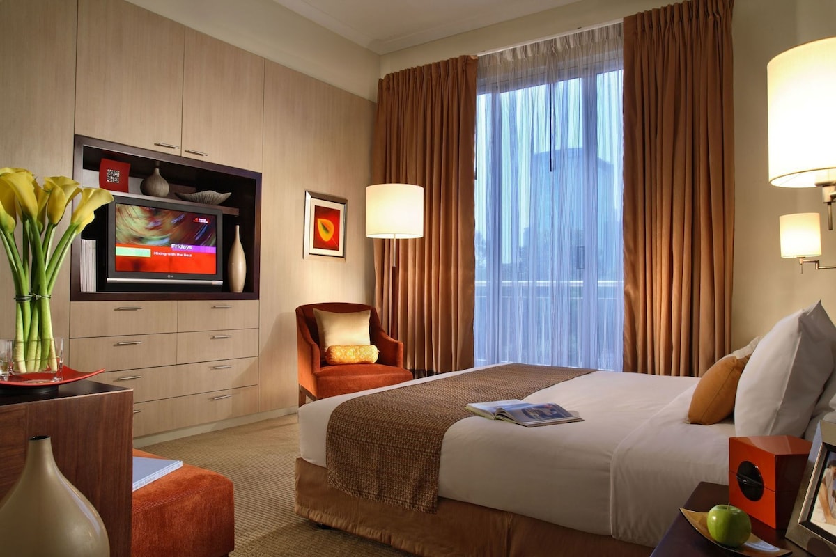 A hotel room with a bed, a TV, a chair, a desk with a vase, and a window with curtains.
