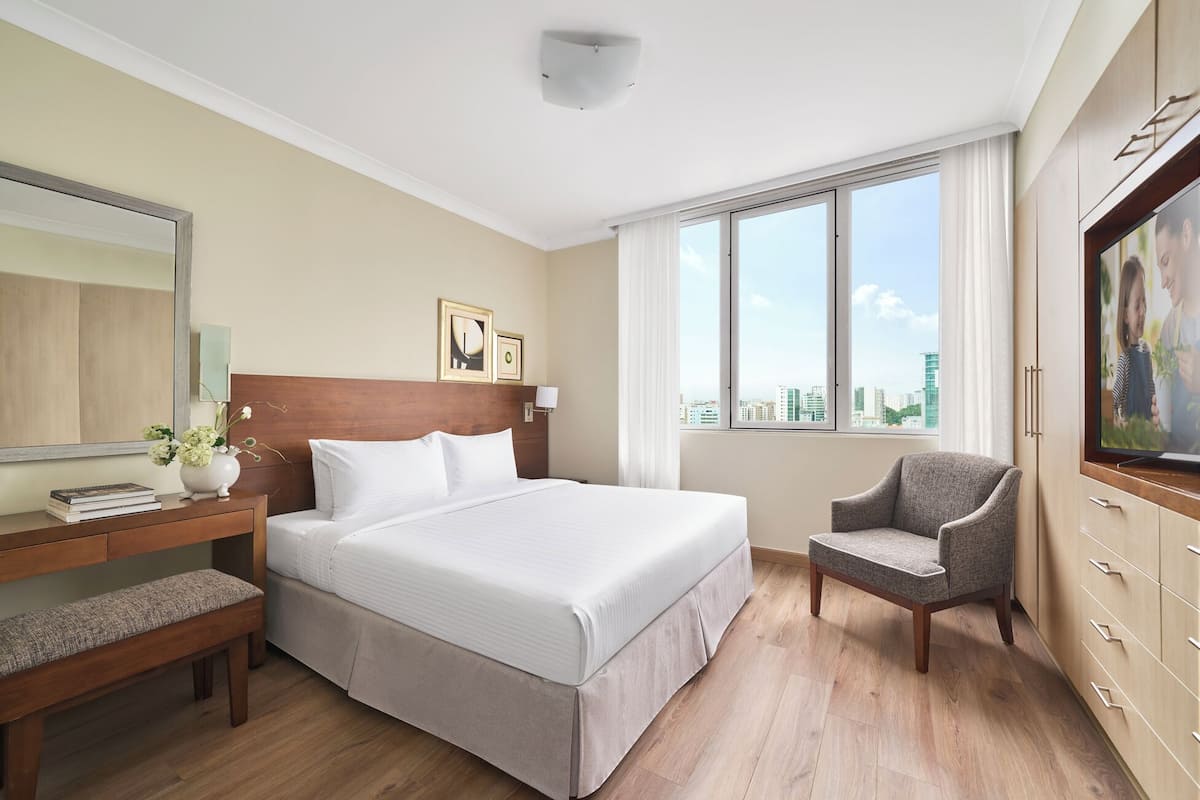A hotel room with a bed, a chair, a desk with a lamp, a mirror, and a window with a city view.