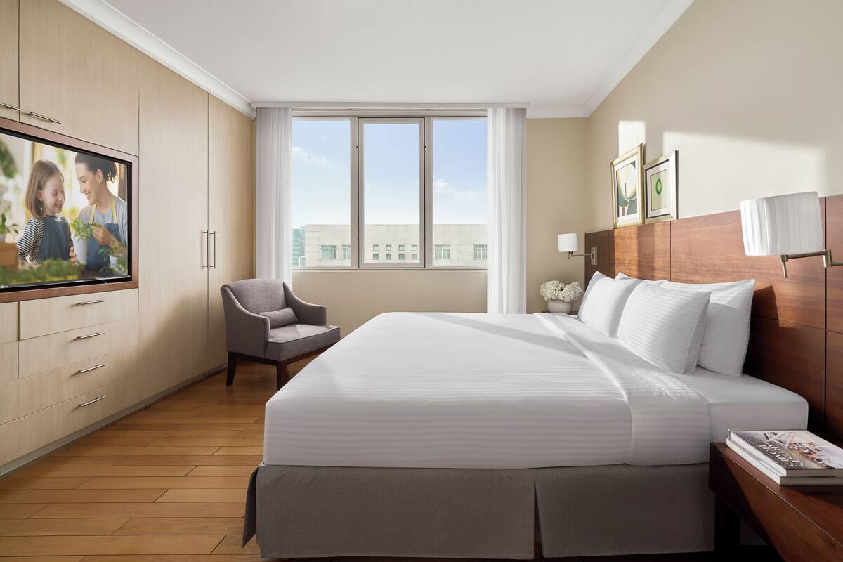A hotel room with a large bed, a TV on the wall, a chair, and a window with a view of buildings.
