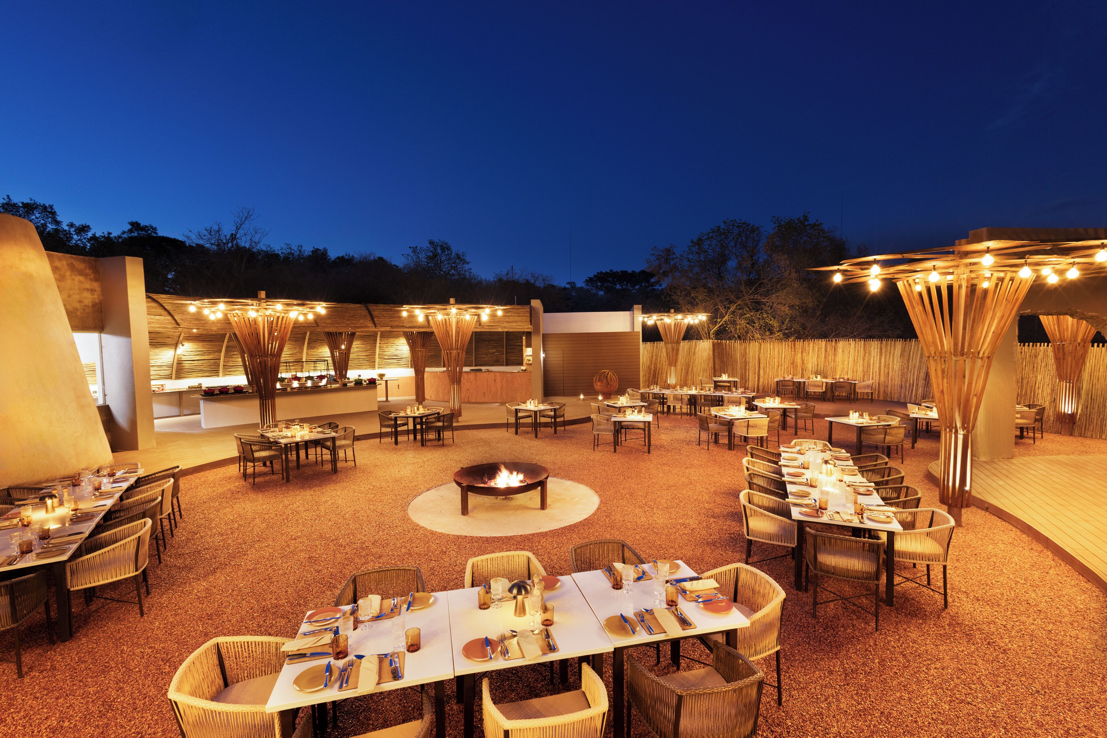 An outdoor dining area with multiple tables and chairs, a central fire pit, and string lights.