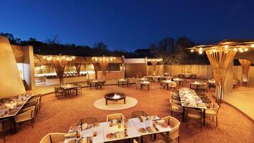 An outdoor dining area with multiple tables and chairs, a central fire pit, and string lights.
