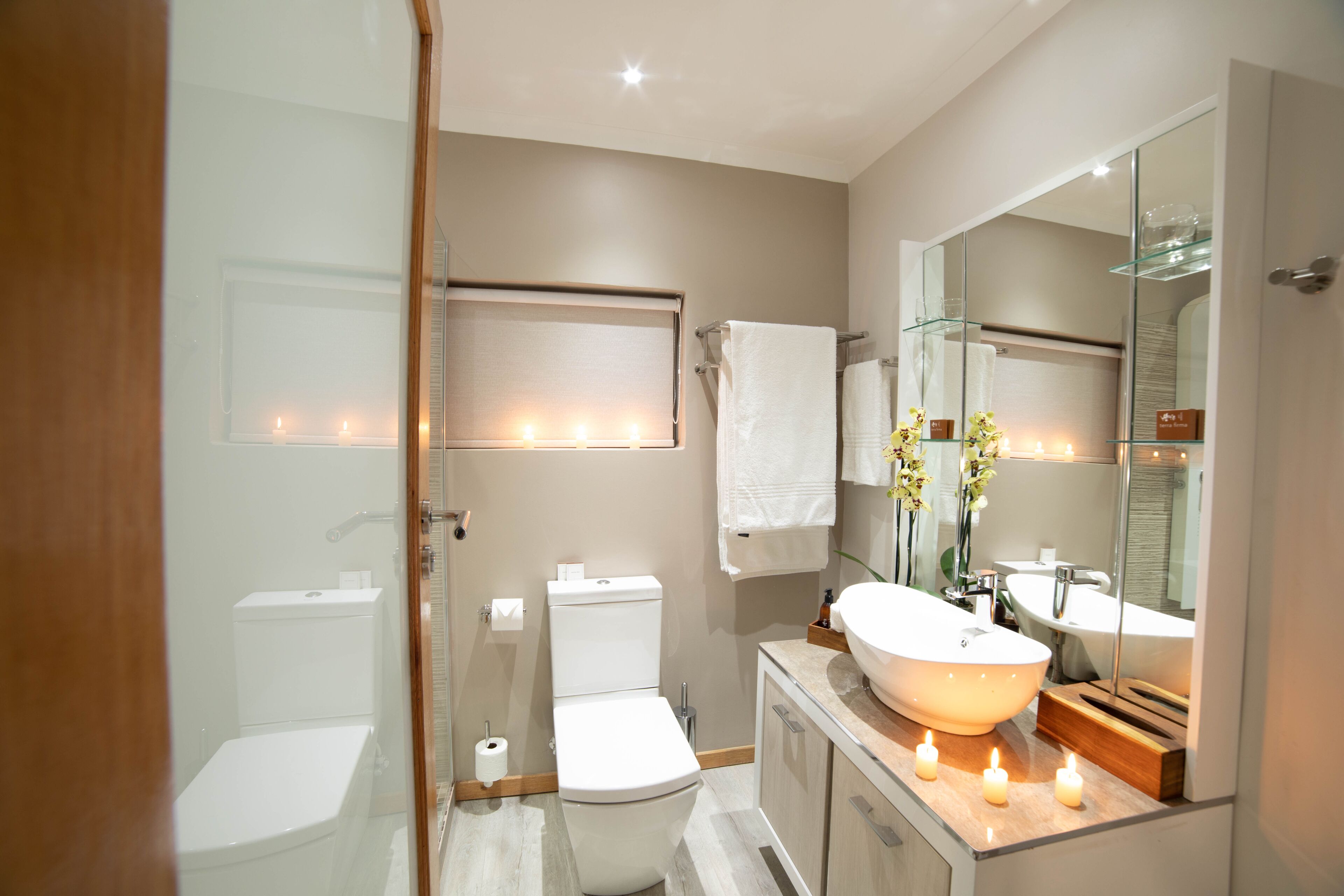 Un baño moderno con cabina de ducha de vidrio, inodoro blanco, tocador con espejo grande y velas en la encimera.