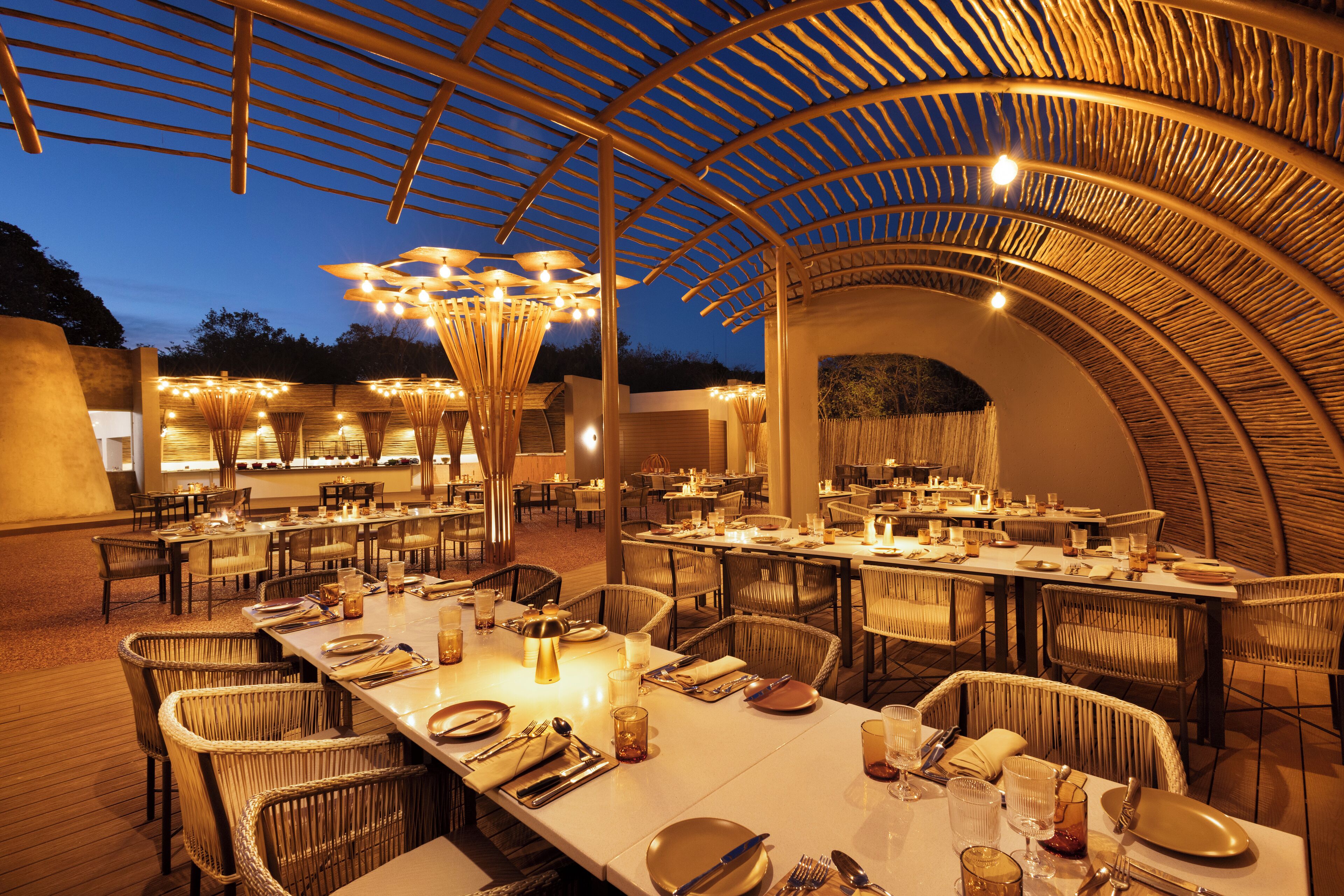 An outdoor dining area with multiple tables and chairs, covered by a wooden pergola with hanging lights.