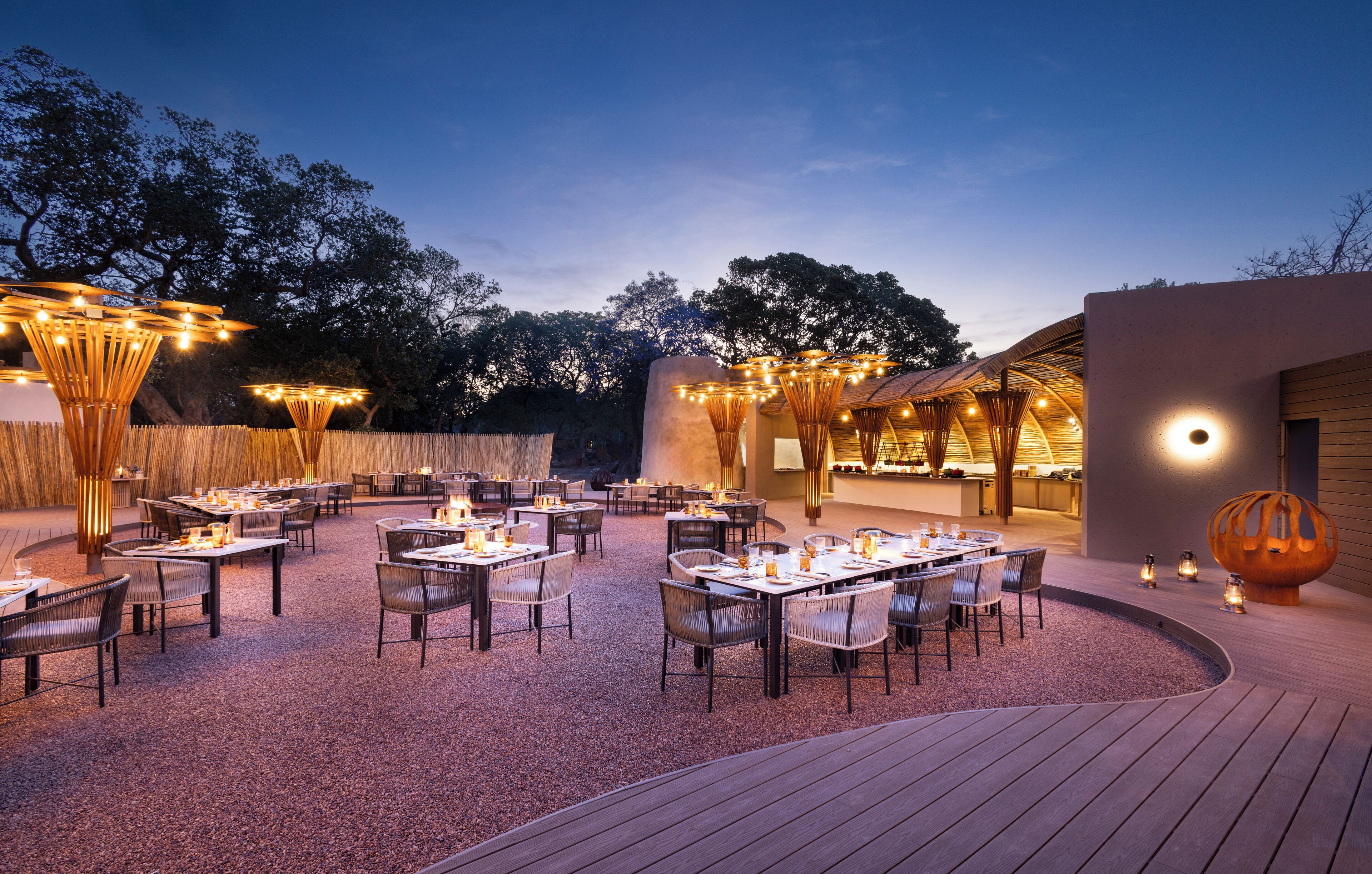 An outdoor dining area with tables and chairs, string lights, and a modern building in the background.
