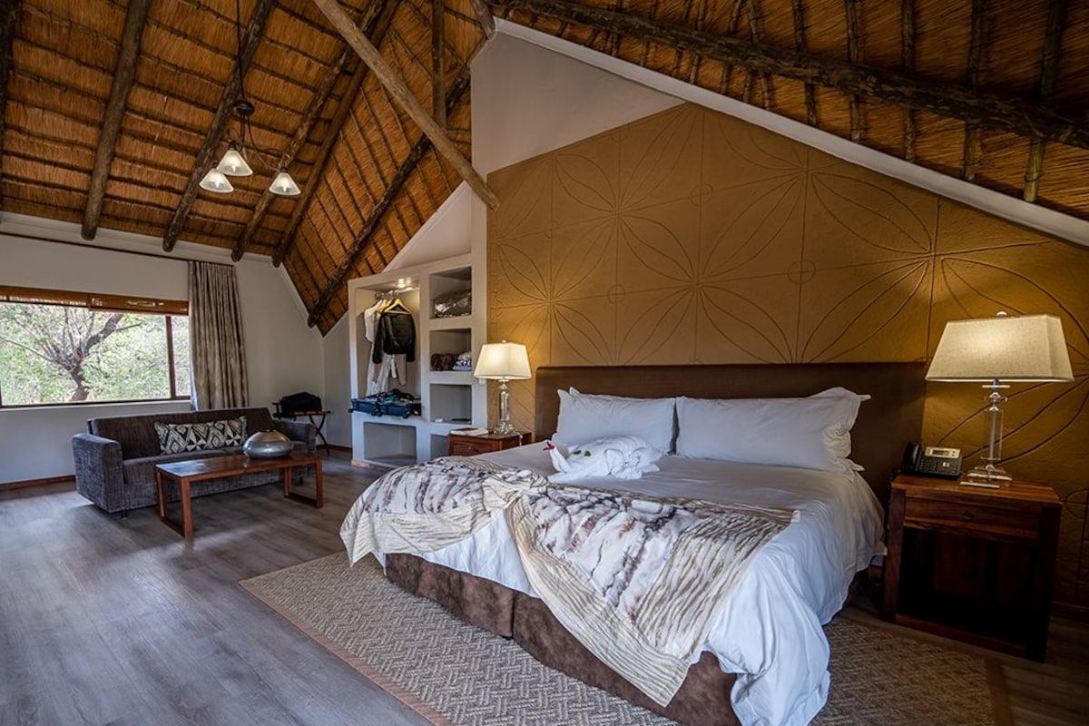 A bedroom with a large bed, two bedside tables with lamps, a wooden headboard, a sofa, and a window with a view of trees.