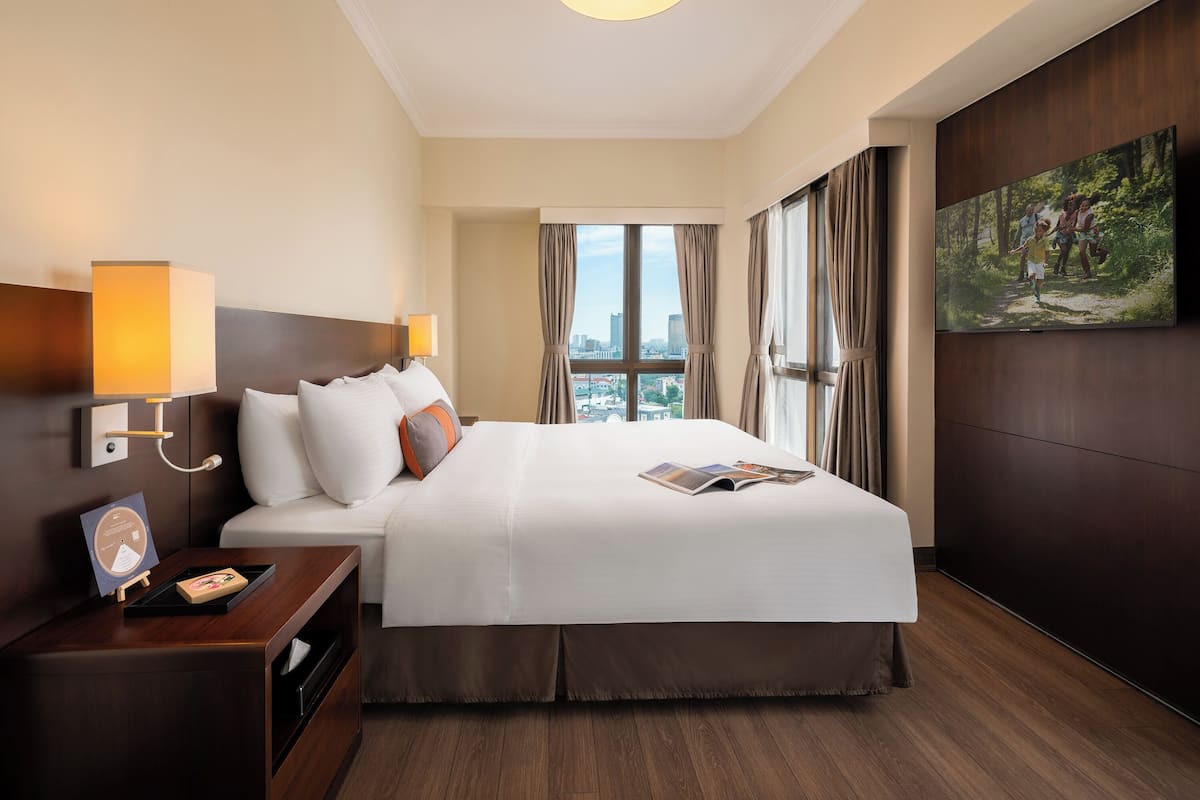 A hotel room with a large bed, a nightstand, a wall-mounted TV, and a city view through the window.