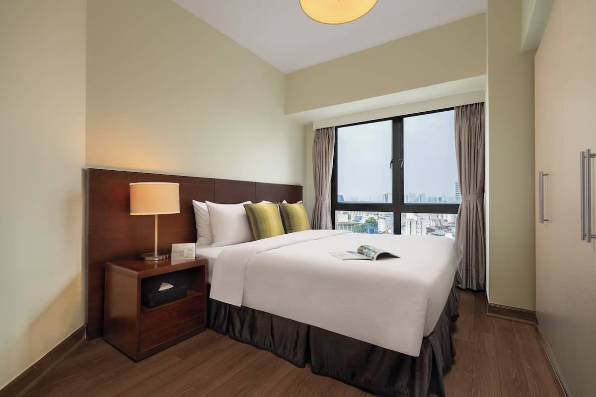 A hotel room with a large bed, a nightstand with a lamp, a window with curtains, and a view of the city.