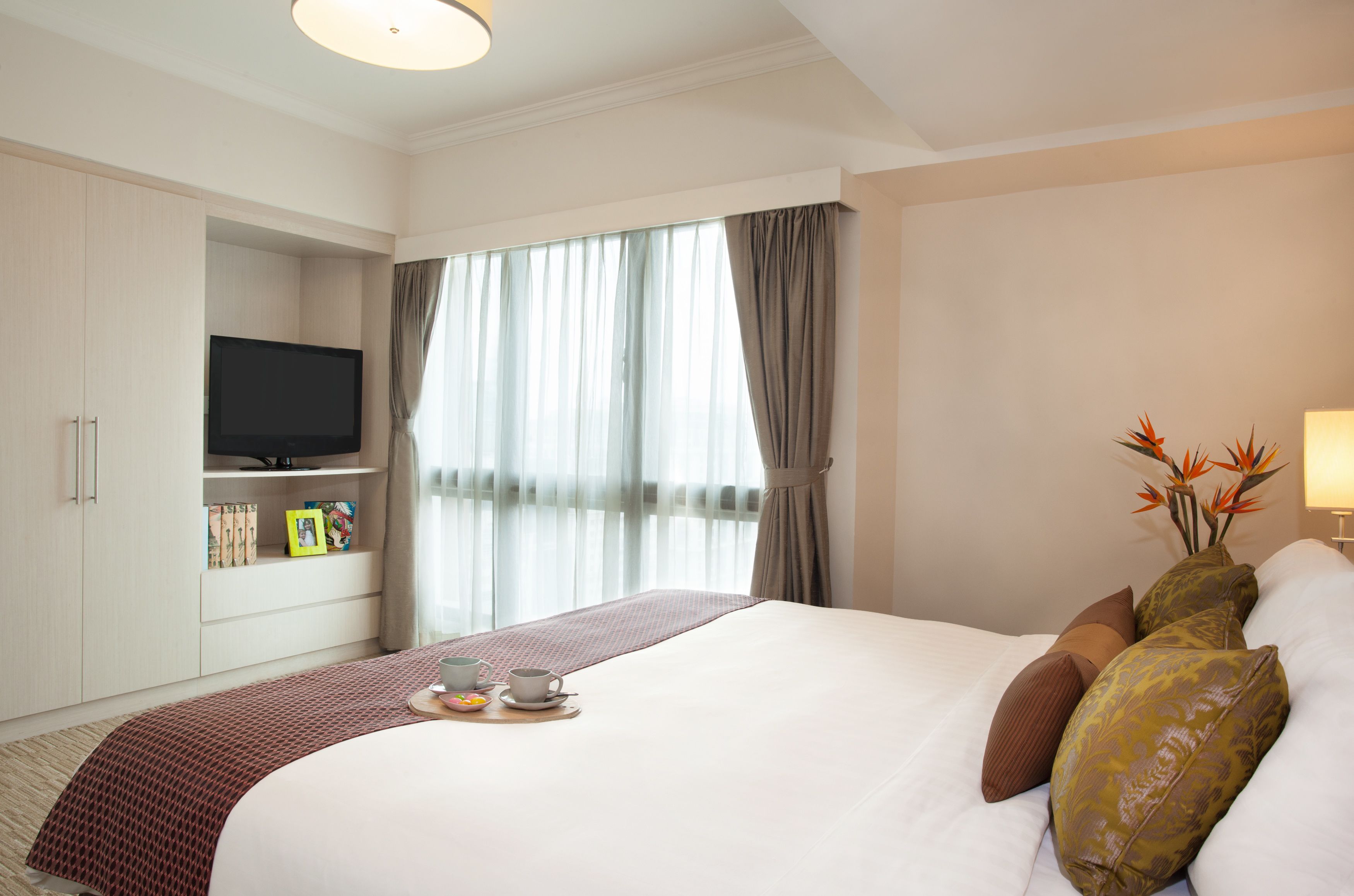 A hotel room with a bed, a television, a window with curtains, and a bedside table with a cup and saucer.