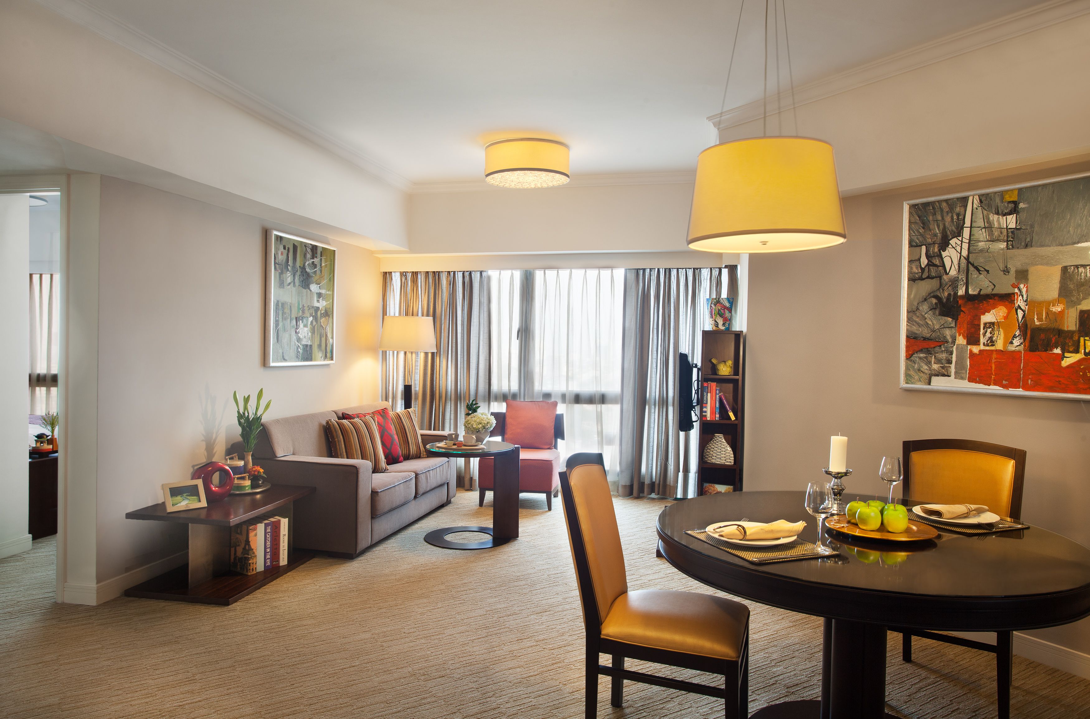 A modern hotel room with a dining area, a sofa, a coffee table, and a bookshelf.