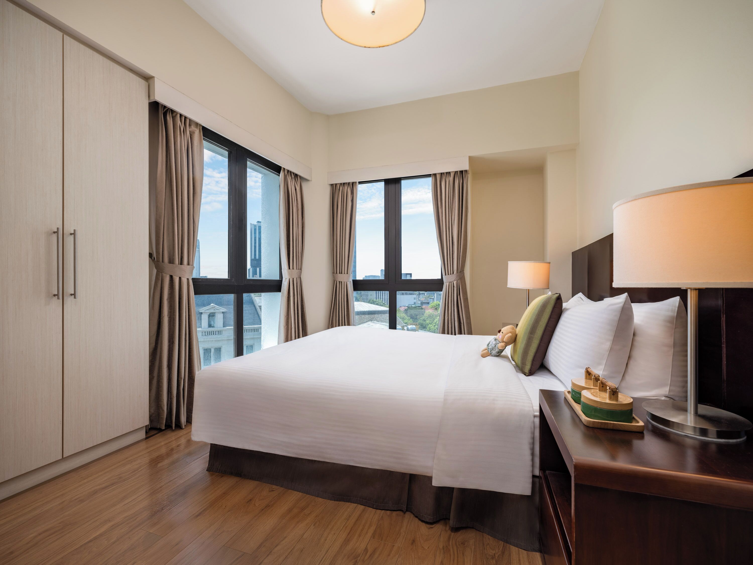 Habitación de hotel con una cama grande, una mesita de noche con lámpara y vistas a la ciudad a través de amplias ventanas.