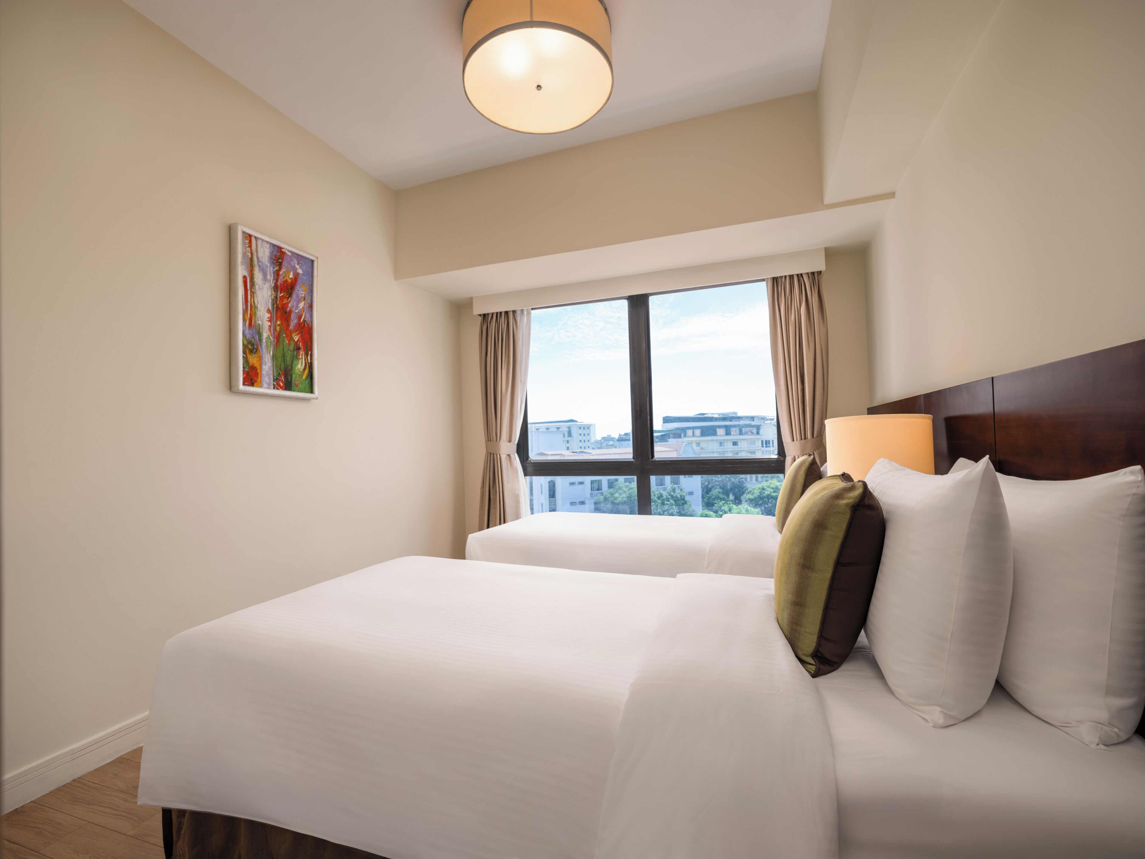 A hotel room with two beds, a large window with curtains, a painting on the wall, and a ceiling light.