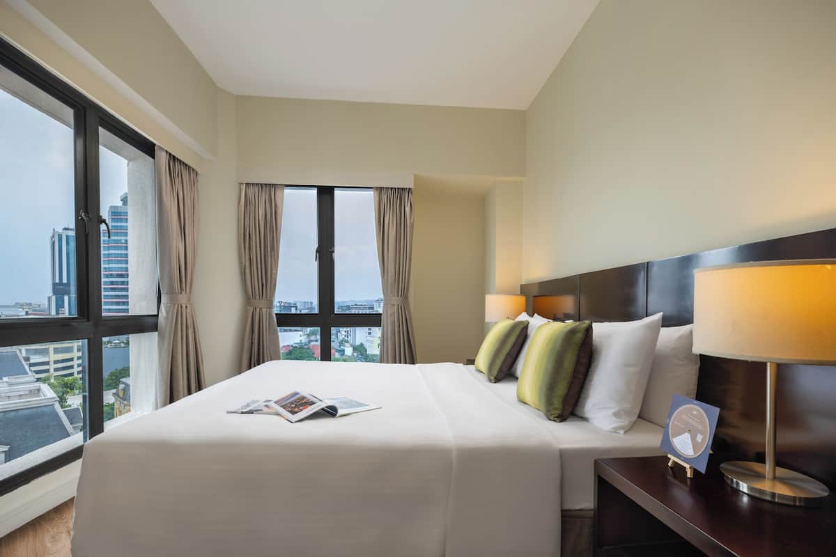 A hotel room with a large bed, two bedside lamps, a nightstand, and a view of the cityscape through large windows.