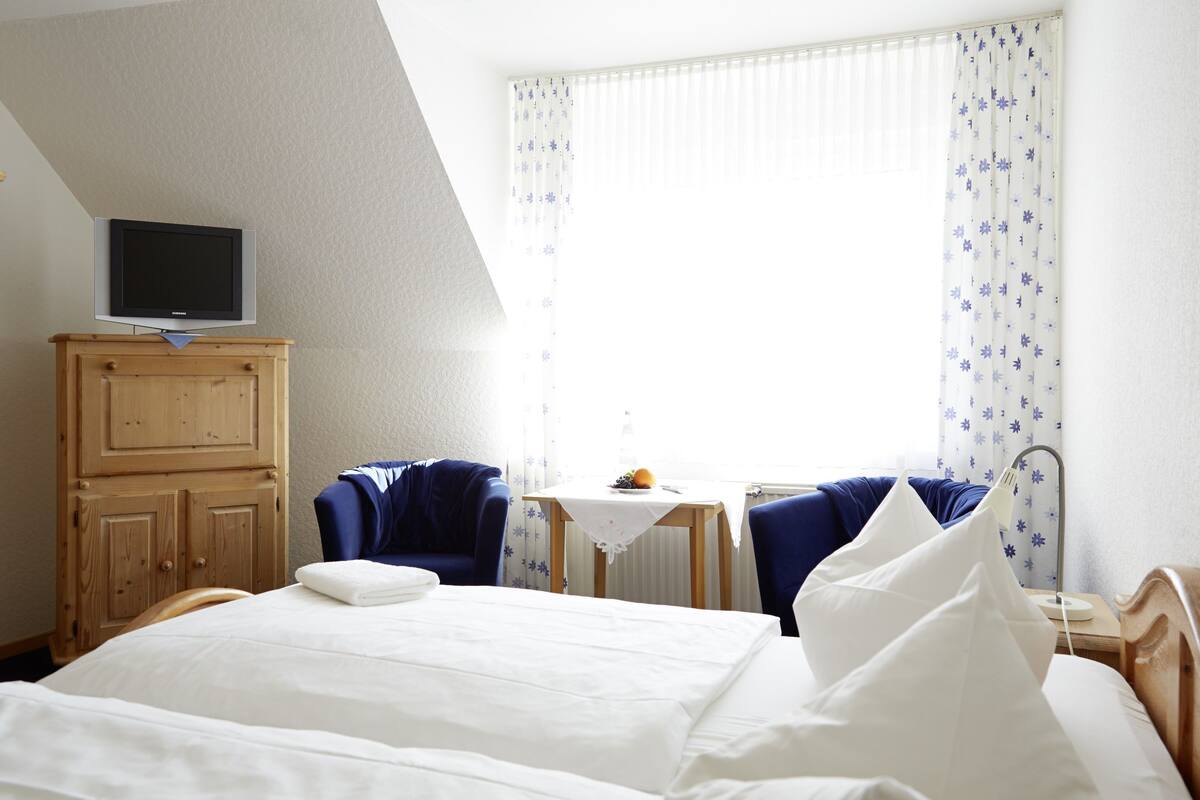 A hotel room with a bed, a television on a wooden cabinet, two chairs, a small table with a fruit bowl, and a window with curtains.