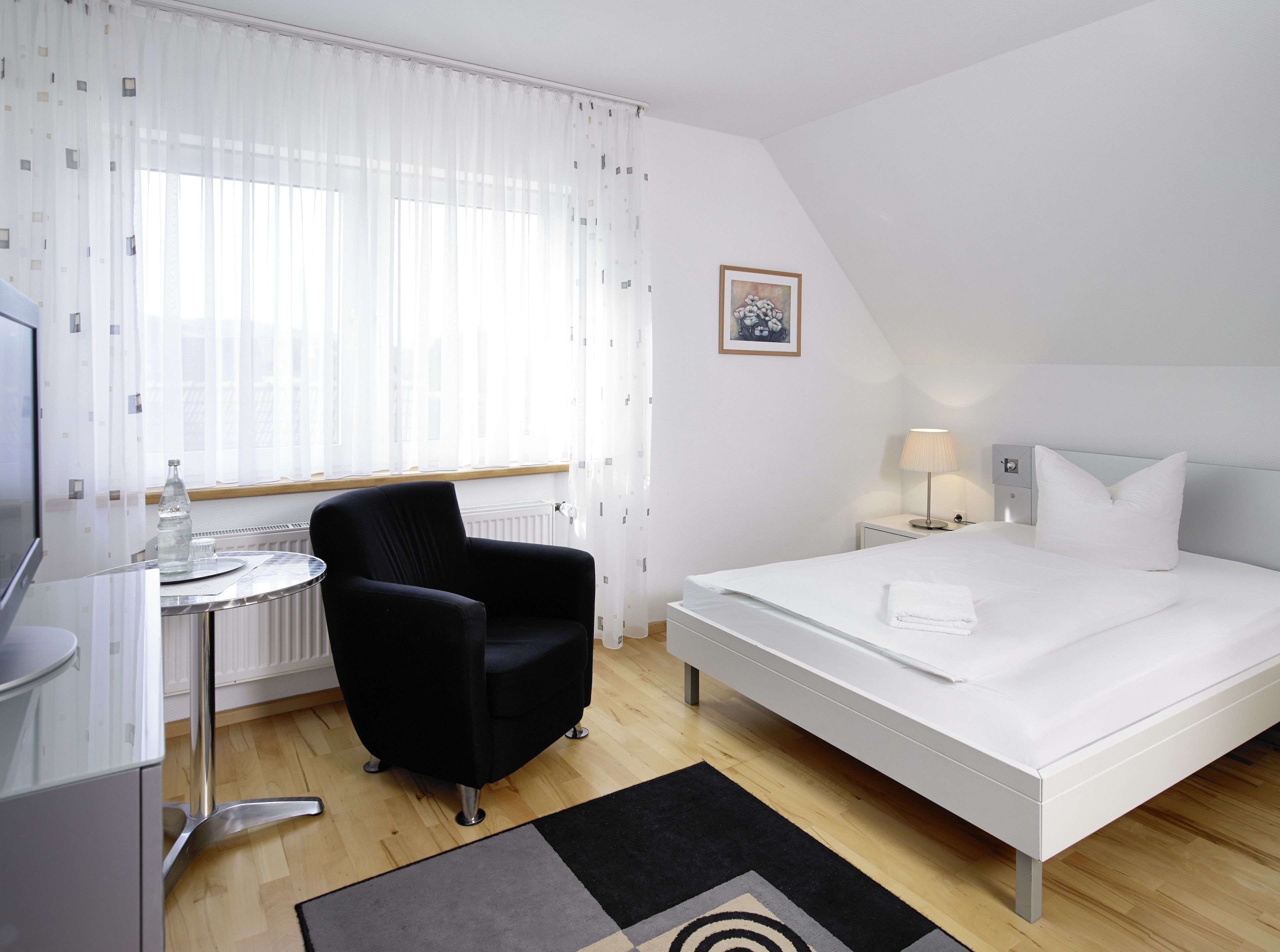 Habitación de hotel con cama, una silla, una mesita, una lámpara y un cuadro en la pared.