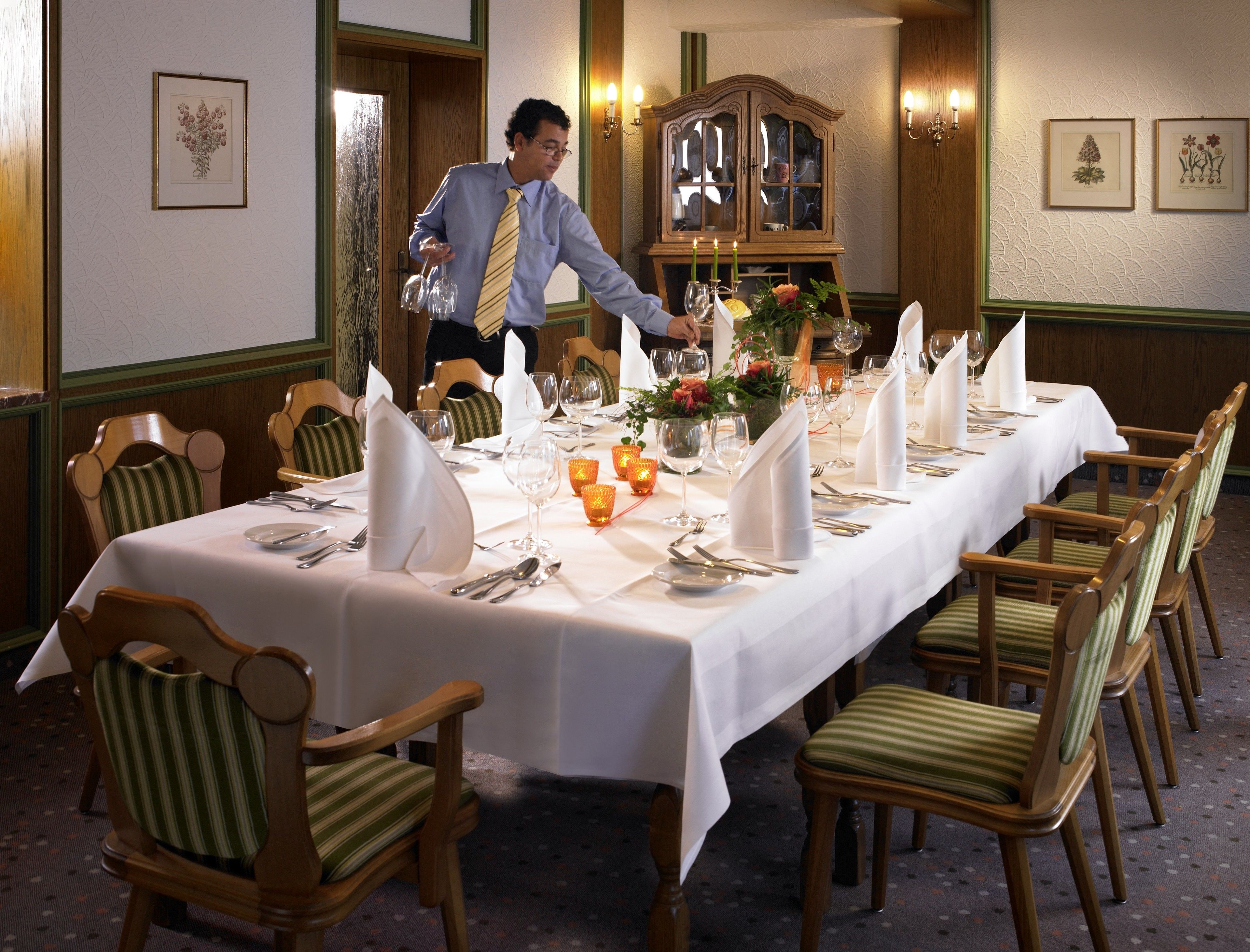 A formal dining room with a long table set for a meal, wooden chairs with striped cushions, a wooden cabinet with glass doors, and wall-mounted sconces.