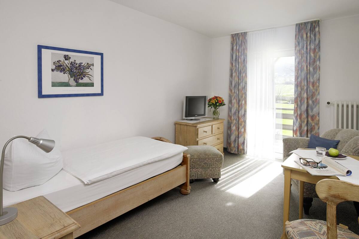 A bedroom with a bed, a desk with a computer, a chair, a lamp, a TV, and a window with curtains.