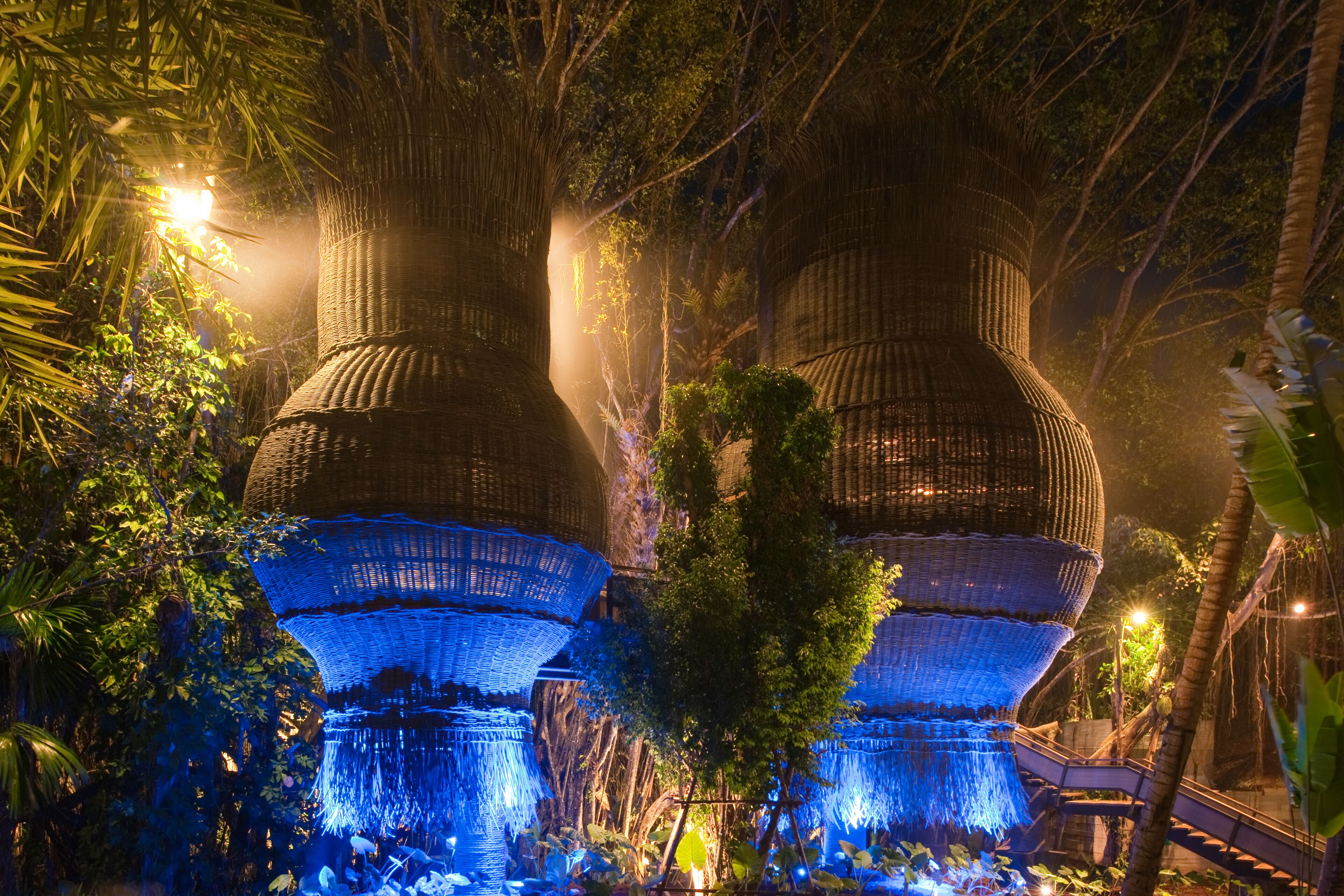 A large, illuminated structure with multiple tiers, surrounded by dense foliage and a pathway leading to it.