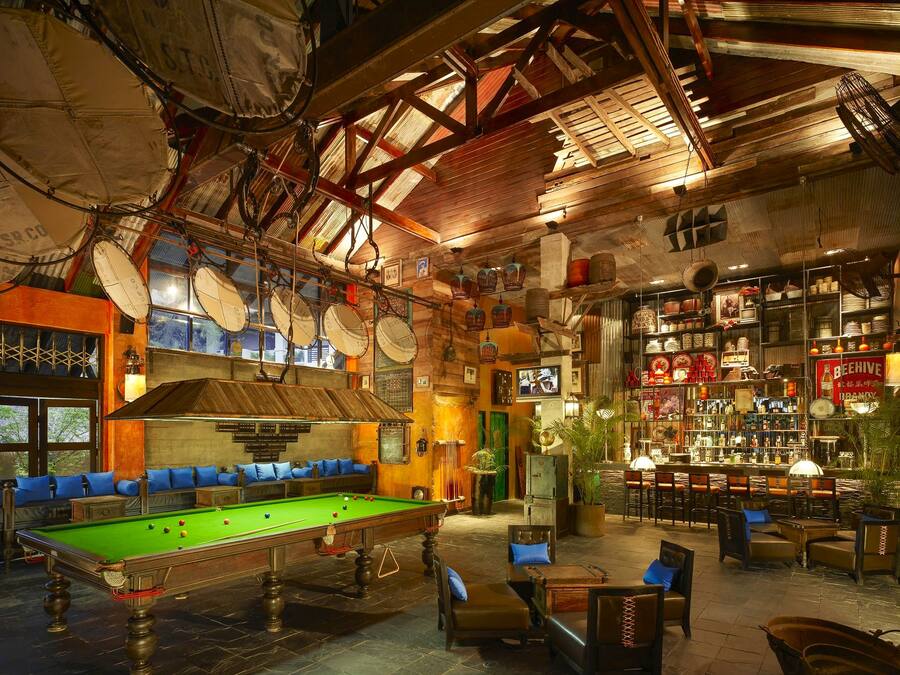 Ein Billardtisch in einem Raum mit Holzbalken und hoher Decke. Es gibt Regale mit verschiedenen Gegenständen sowie eine Bar mit Stühlen und Tischen.