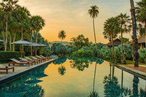 A pool with lounge chairs and umbrellas, surrounded by palm trees and a building in the background. - The Slate (Sa Khu)