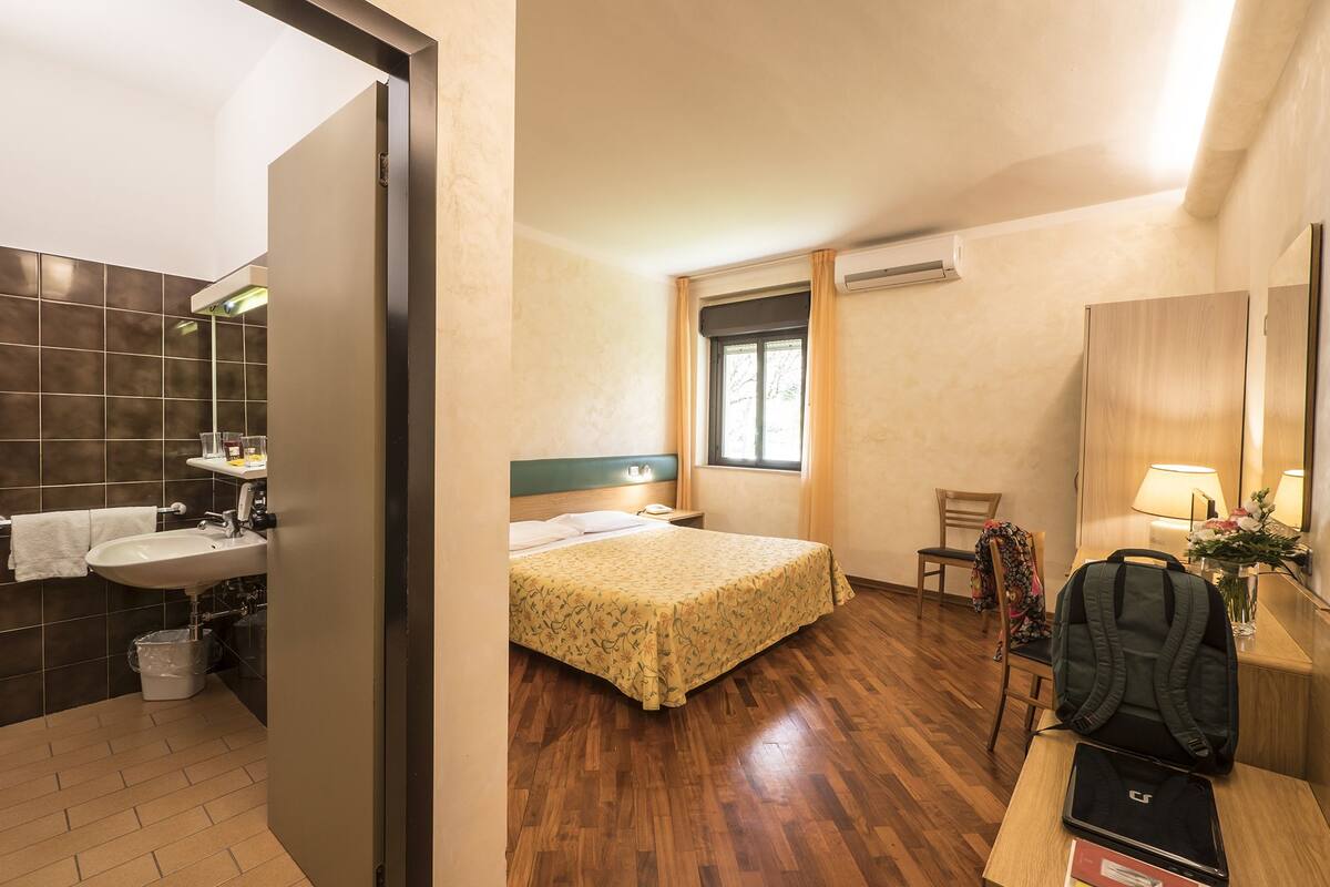 Une chambre d’hôtel comprenant un lit, un bureau avec un ordinateur portable, une chaise, une petite table avec une lampe, une fenêtre et une salle de bain avec un lavabo et un miroir.