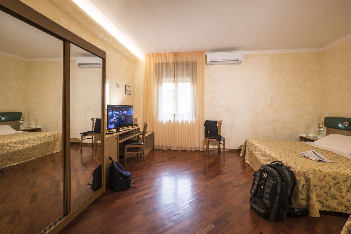 Une chambre d’hôtel avec un plancher en bois, un lit recouvert d’une courtepointe à motifs floraux, un bureau avec une télévision, une chaise, un miroir et un appareil de climatisation.
