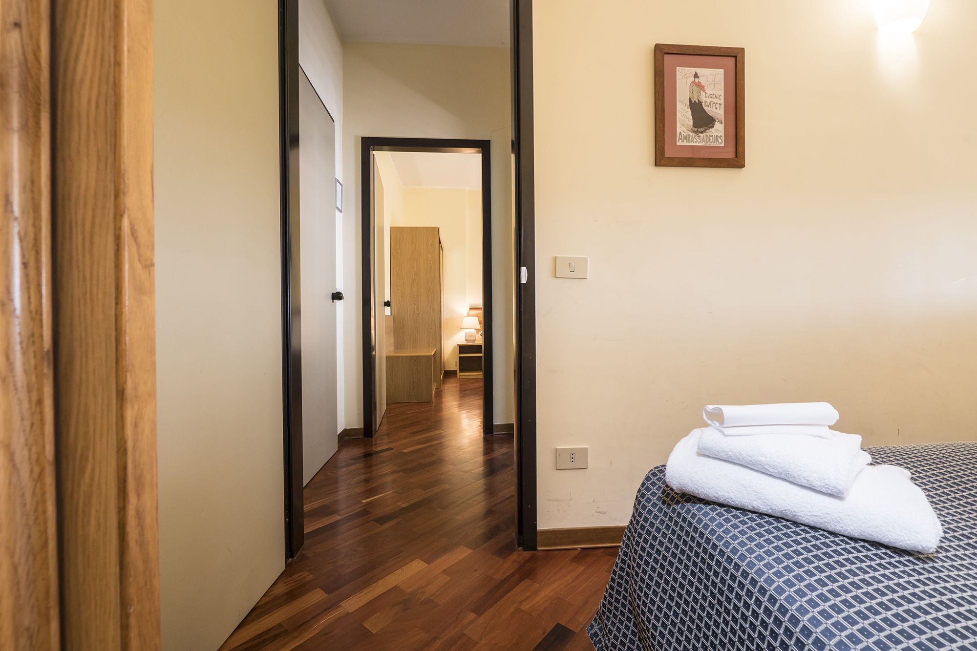 A hotel room with a wooden floor, a bed with a patterned cover, a framed picture on the wall, and a hallway leading to another room.