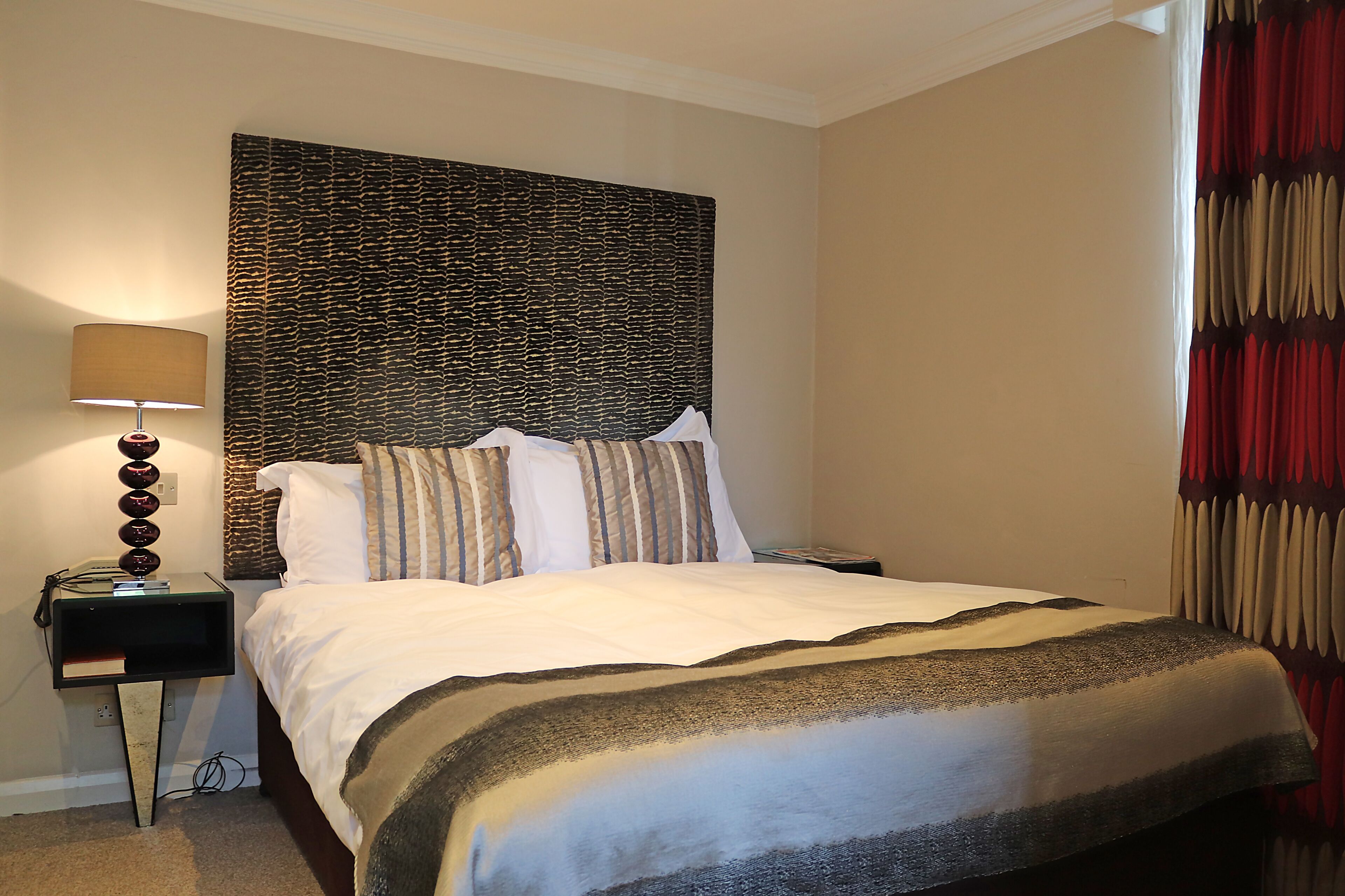 A neatly made bed with a patterned headboard, a bedside lamp, and a nightstand with a telephone and a book.