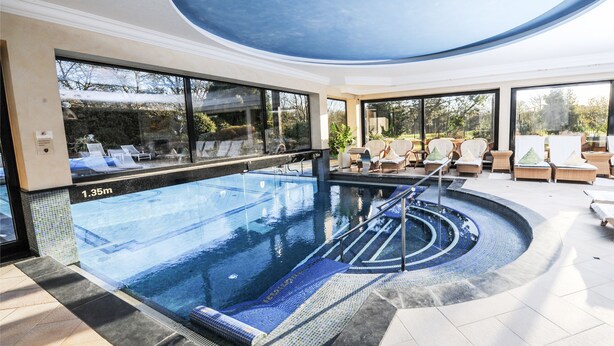 Una piscina interior moderna con agua azul cristalina, sillas de descanso y vistas al exterior.