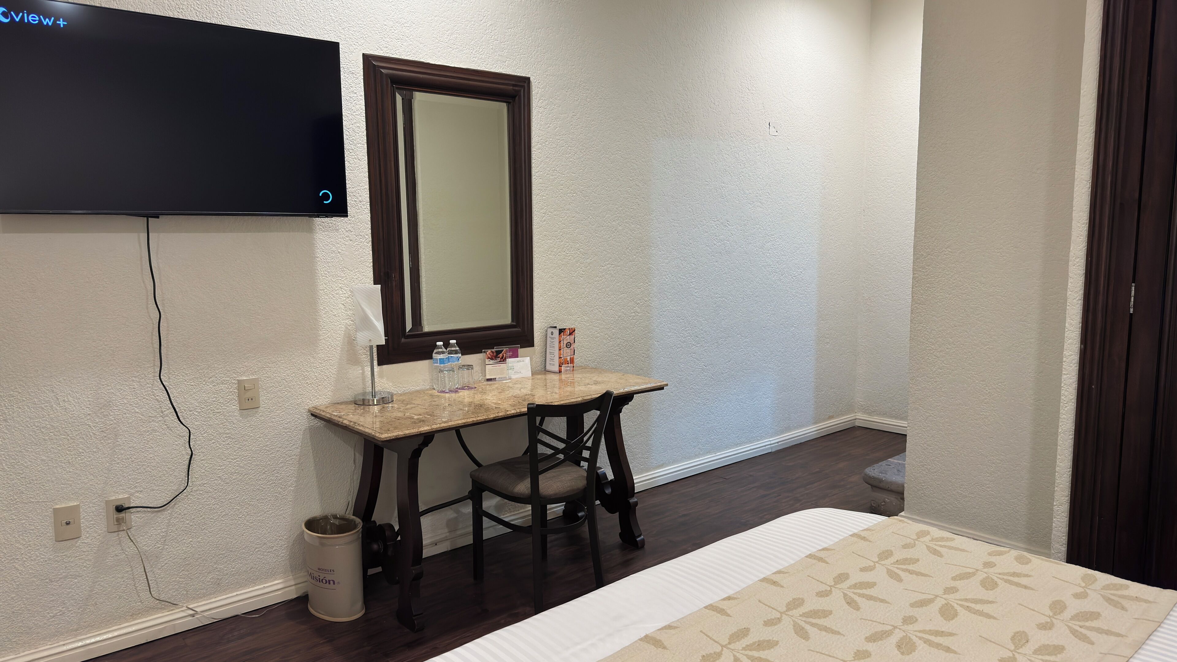 Habitación de hotel con televisión de pantalla plana montada en la pared, un escritorio de madera con una silla, un cesto de basura pequeño y una cama con cubrecama estampado.