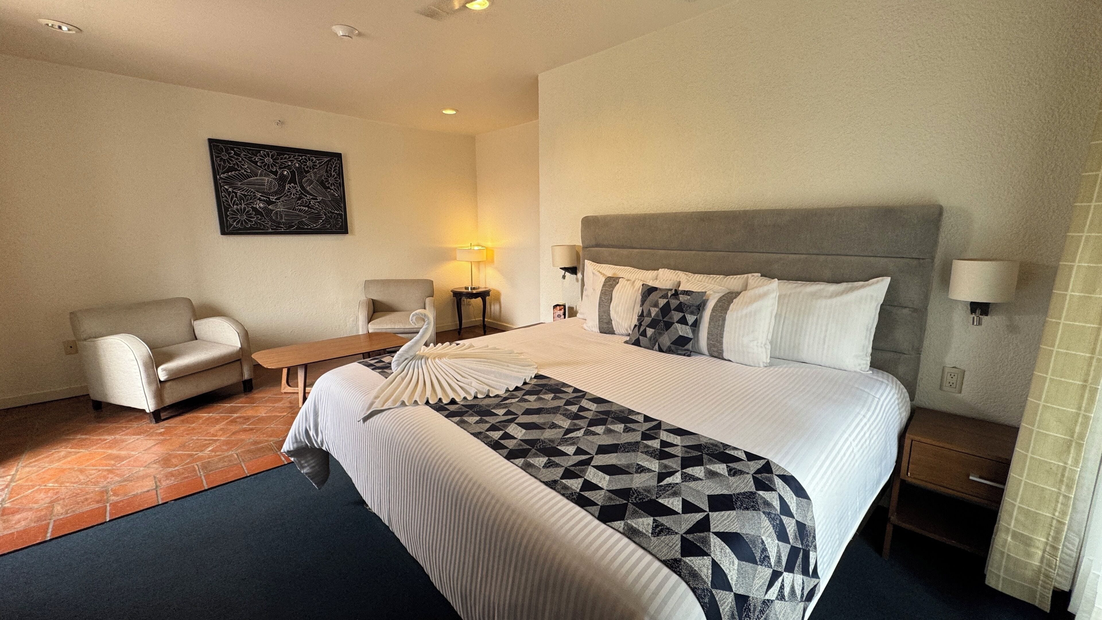 Habitación de hotel con una cama, dos sillones, una mesita y una lámpara de pared, además de un cuadro.