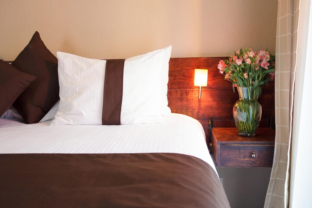A neatly made bed with white and brown bedding, a wooden nightstand with a vase of flowers, and a wall-mounted lamp.