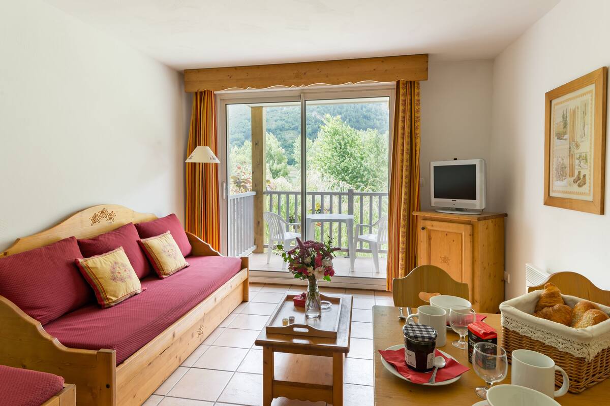 A cozy room with a wooden sofa, a dining table with a vase of flowers, a television, and a basket of bread.