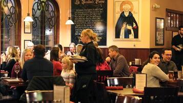 En restaurant med gæster ved borde, en tjener der serverer mad, og en væg med religiøse kunstværker.