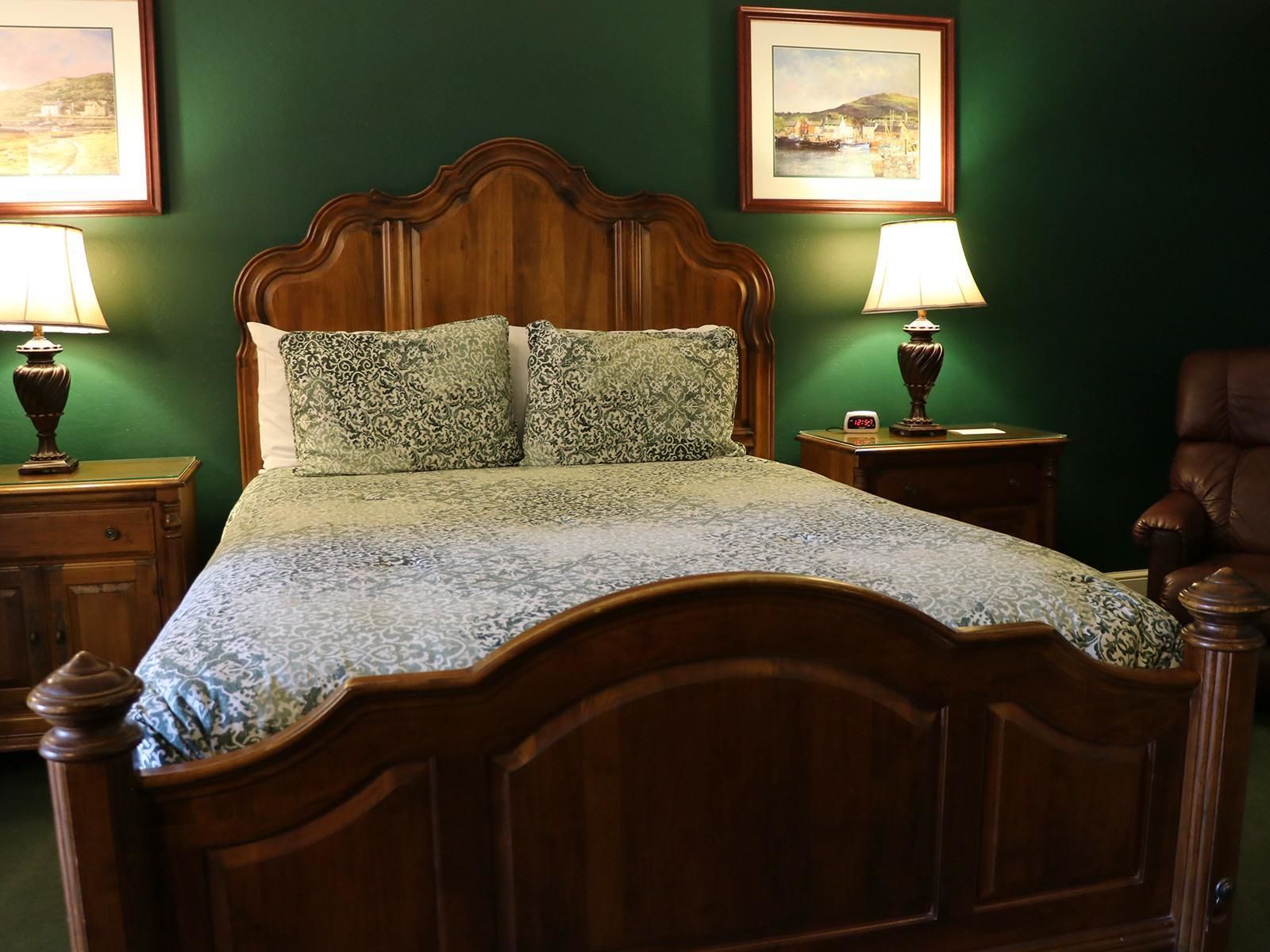 Una cama con dosel de madera con un edredón estampado, flanqueada por dos mesitas de noche con lámparas, frente a una pared verde con cuadros enmarcados.
