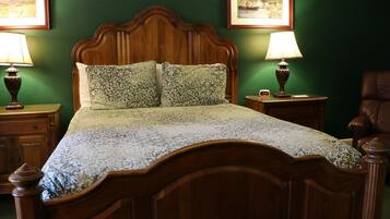 Una cama con dosel de madera con un edredón estampado, flanqueada por dos mesitas de noche con lámparas, frente a una pared verde con cuadros enmarcados.