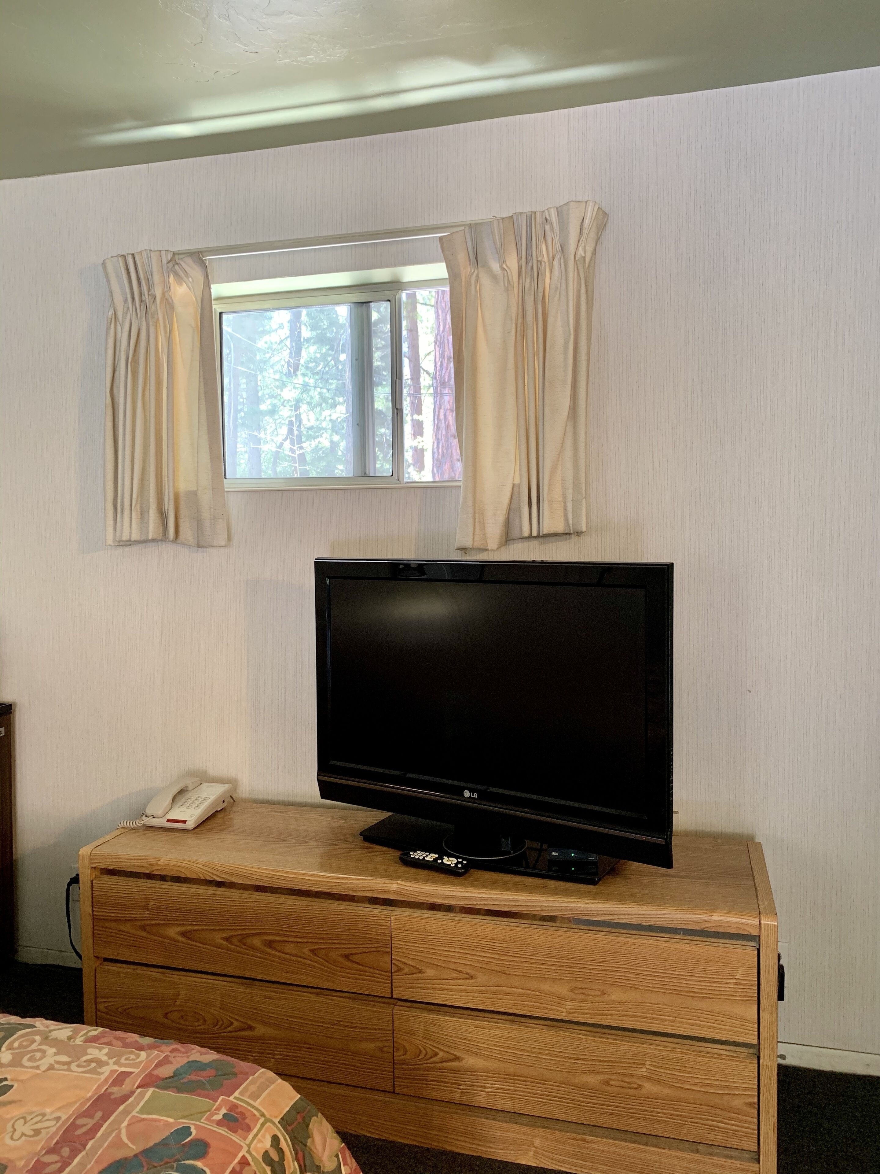 A room with a wooden dresser, a television on top, a telephone, and a window with curtains.