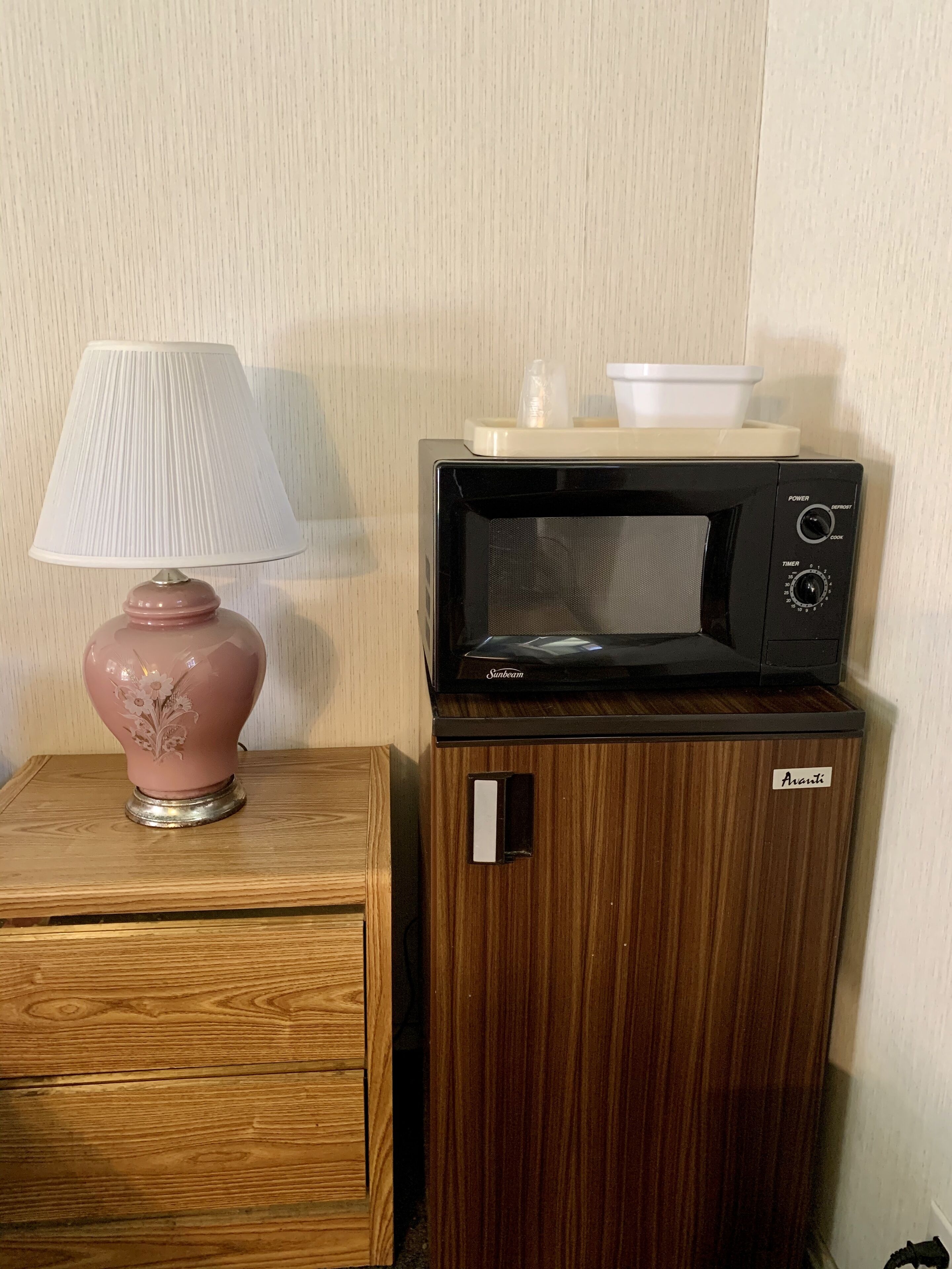 A compact microwave oven with a control panel, a small wooden cabinet, and a pink lamp with a floral design on a wooden table.