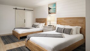 A hotel room with two wooden beds, white bedding, a wooden headboard, a nightstand with a lamp, a painting of a beach, and a sliding door.