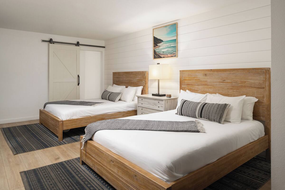 A hotel room with two wooden beds, white bedding, a wooden headboard, a nightstand with a lamp, a painting of a beach, and a sliding door.