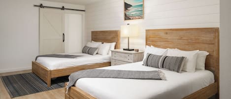 A hotel room with two wooden beds, white bedding, a wooden headboard, a nightstand with a lamp, a painting of a beach, and a sliding door.