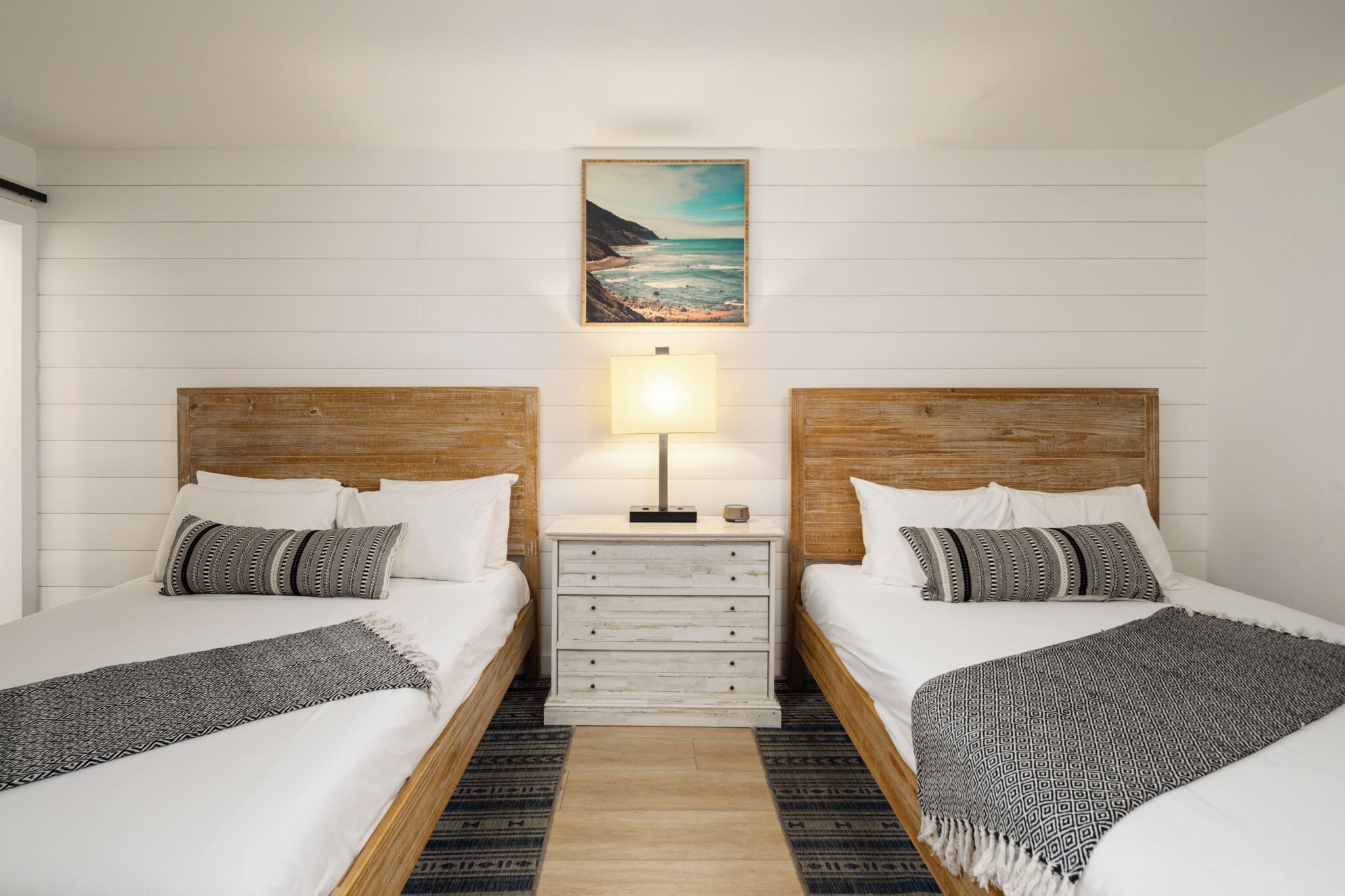 A double room with two beds, a nightstand with a lamp, and a painting of a beach scene.