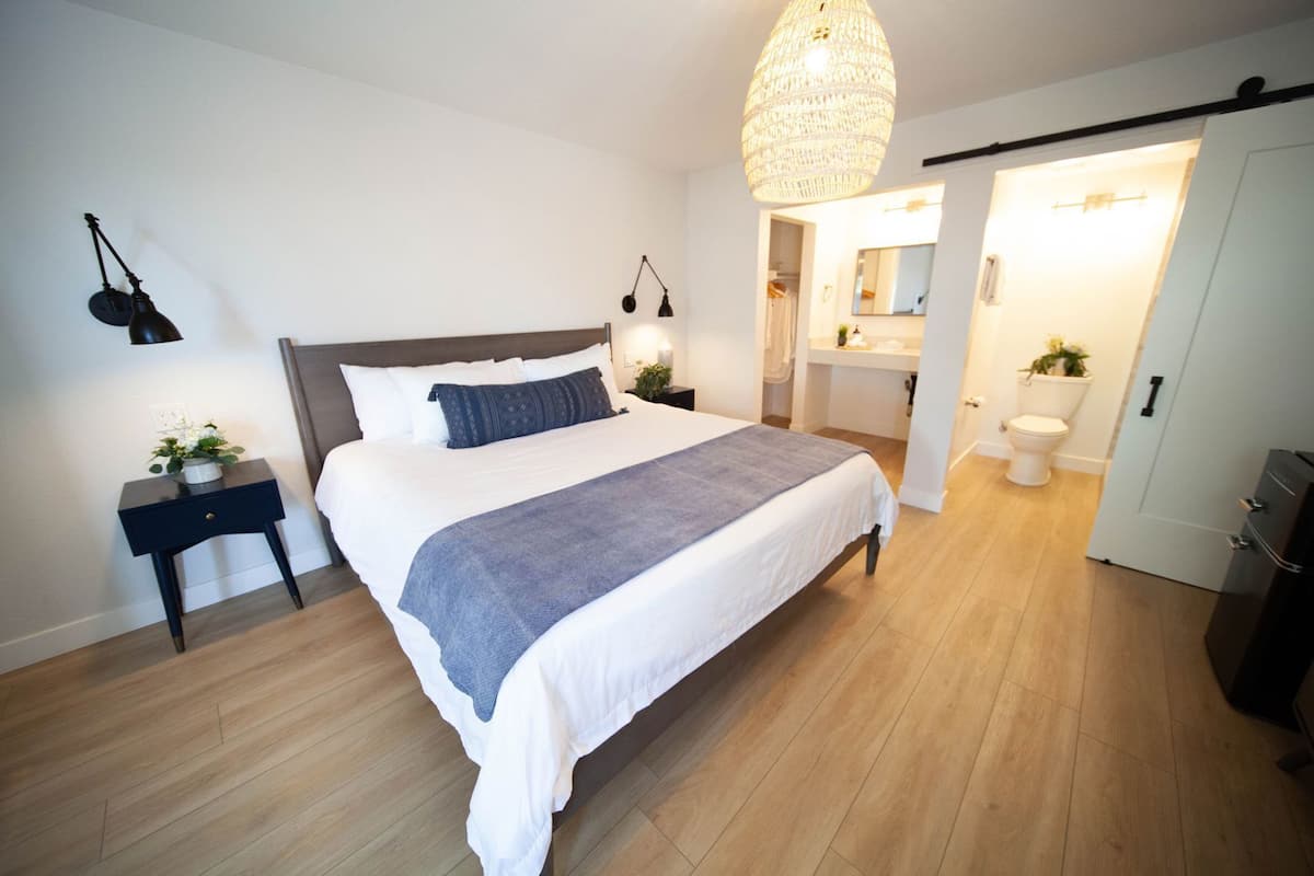 A modern bedroom with a large bed, bedside lamps, a hanging light fixture, and a view into the bathroom.