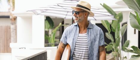 Ein Mann geht mit einem Surfbrett auf einem Balkon spazieren, er trägt einen Hut, eine Sonnenbrille und Freizeitkleidung.