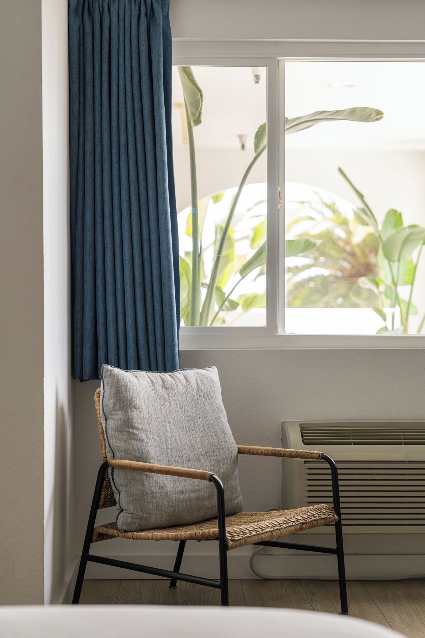 Ein Zimmer mit einem Korbstuhl, einem blauen Vorhang und einem Fenster, durch das Pflanzen draußen sichtbar sind.